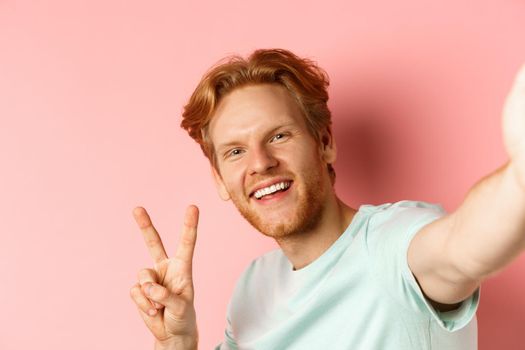 Close-up of happy redhead man taking selfie on smartphone and showing peace sign, view from phone camera, pink background.