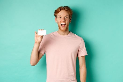 Cheerful young man with red hair and beard, wearing t-shirt, showing plastic credit card and smiling at camera, demonstrate new bank promo.
