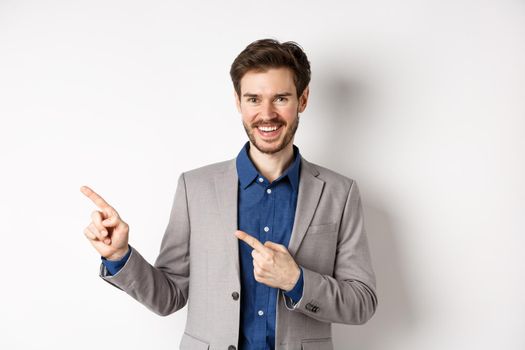 Handsome caucasian man in business suit pointing fingers left at banner and smiling, showing advertisement, standing against white background.