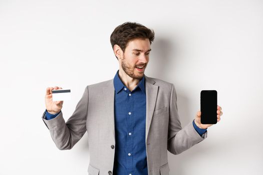 Online shopping. Smiling business man in suit showing plastic credit card with empty smartphone screen, standing against white background.