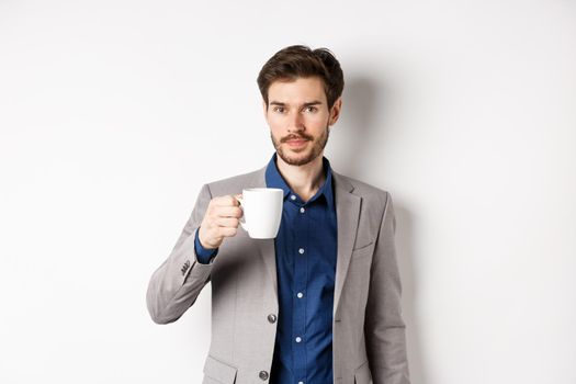 Handsome businessman in suit drinking coffee or tea from office mug, standing against white background.