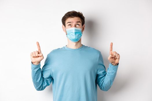 Coronavirus, health and quarantine concept. Curious guy reading banner on top, looking and pointing fingers up, standing in medical mask over white background.