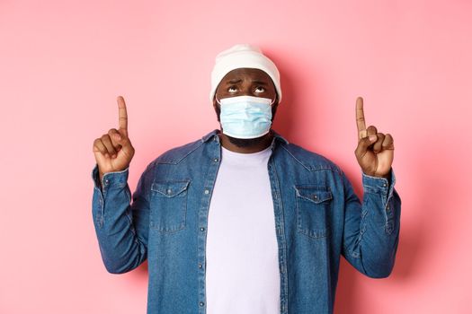Coronavirus, lifestyle and global pandemic concept. Worried and anxious african-american man in face mask, pointing and looking up concerned, standing over pink background.
