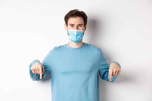 Covid-19, health and quarantine concept. Smiling caucasian man in face mask pointing fingers down, showing logo, standing over white background.
