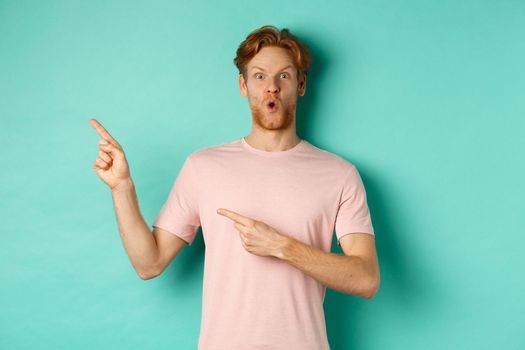 Amazed young man with red hair and beard, gasping amazed, pointing at upper left corner and showing promo, checking out advertisement, standing over turquoise background.