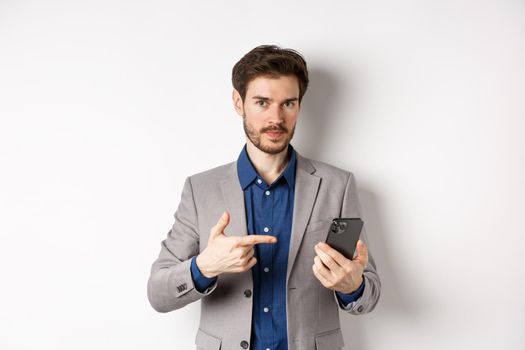 Caucasian business man in suit pointing at smartphone, showing on mobile phone, white background.