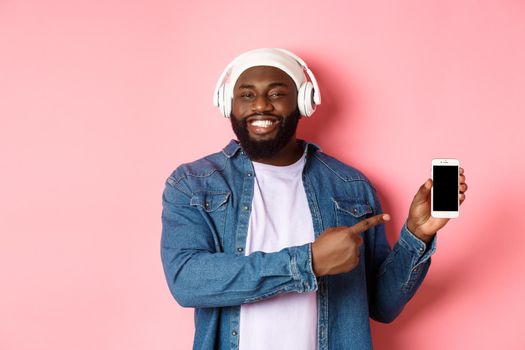 Happy black man listening music in headphones and smiling, pointing mobile phone screen app or playlist, standing over pink background.
