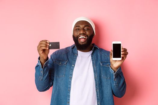 Online shopping. Happy Black man laughing and smiling, showing credit card and smartphone screen, recommending phone app, pink background.