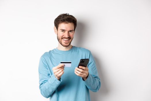 E-commerce and shopping concept. Smiling young man making online payment with plastic credit card and smartphone, standing over white background.