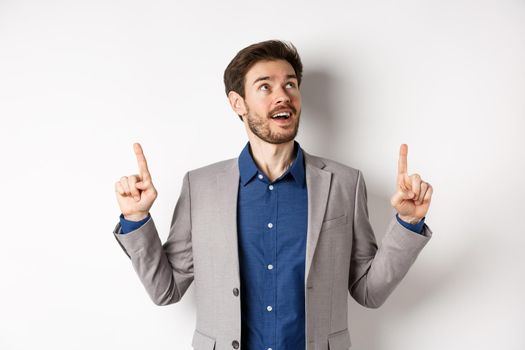 Excited handsome man with beard, wearing suit and looking amused up, pointing at top with admiration, standing against white background.