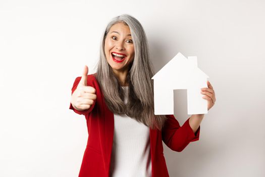 Real estate. Portrait of asian female broker showing thumb-up and paper house cutout, recommending agency to buy property, standing happy over white background.