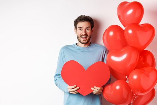 Smiling boyfriend holding Valentines card and standing near romantic red heart balloons, celebrating lovers day, standing over white background.