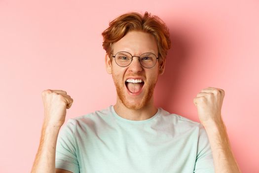 Close up of satisfied lucky redhead man winning, shouting from joy and making fist pump, celebratig victory, standing like champion over pink background.