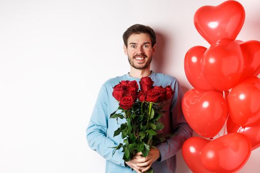 Valentines day romance. Excited young man with bouquet of red roses and heart balloons smiling at camera, bring presents for lover on valentine date, standing over white background.