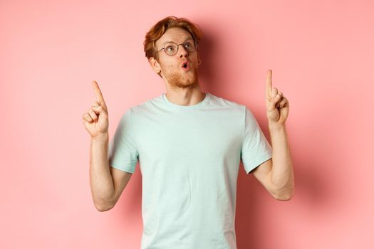 Wow check this out. Surprised redhead guy in glasses looking and pointing up, gasping amazed, standing over pink background.