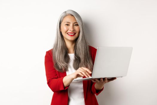 Business. Senior woman working on laptop, wearing office outfit and smiling, standing over white background.