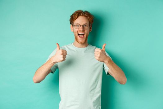 Excited young man with red hair, wearing glasses, showing thumbs-up and agree or praise something, smiling amazed and saying yes, standing over turquoise background.