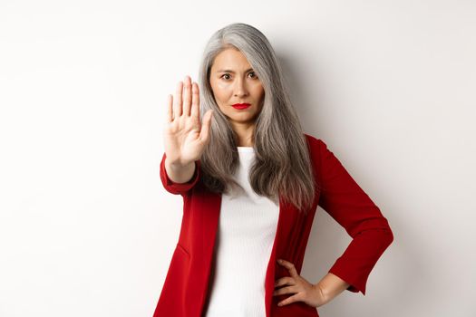 Serious asian woman in red blazer showing stop sign, telling no, frowning and rejecting something bad, standing against white background.
