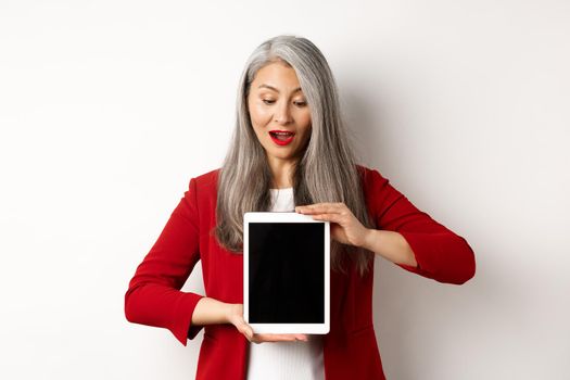 Business. Mature asian office lady showing digital tablet screen and looking down amazed, standing over white background.