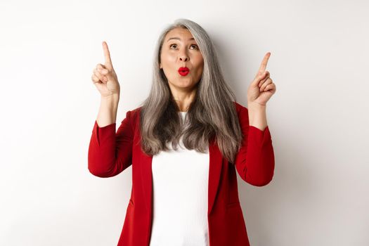 Amazed korean businesswoman with grey hair, wearing red blazer at work, pointing fingers up and looking surprised, standing over white background.