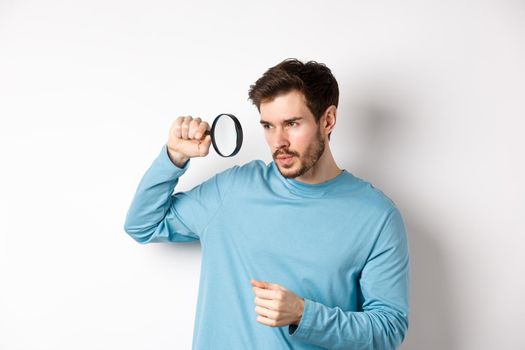 Young handsome man look through magnifying glass with curious face, investigating or searching for something, standing on white background.