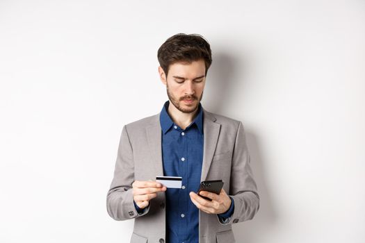 Handsome businessman paying online with credit card and smartphone, standing on white background in suit.