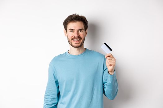 Smiling male bank client showing plastic credit card with happy face, standing over white background.