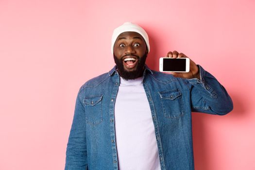 Online shopping and technology concept. Amazed Black man showing phone screen horizontally and staring at camera excited, standing over pink background.