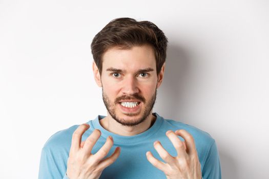 Close up of angry young man with beard, shaking hands mad, squeeze teeth and frowning furious, standing outraged over white background.