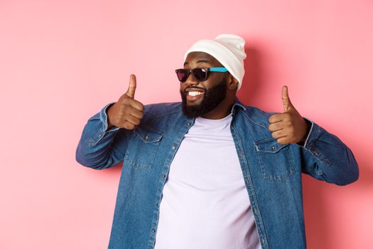 Happy and satisfied african-american guy in sunglasses showing thumbs-up, praise and like promo, agree with you, standing over pink background.