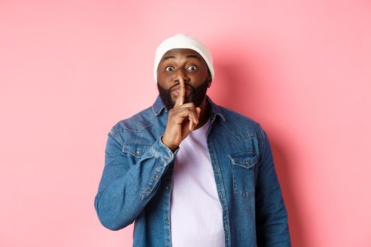 Handsome young black guy in beanie and denim shirt prepare surprise, hushing softly and looking at camera, asking keep quiet, pink background.