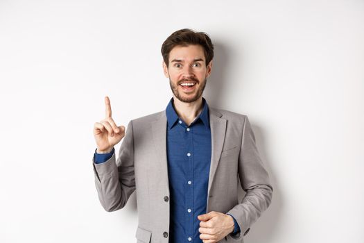 Excited businessman in suit pitching good idea, raising finger in eureka sign and looking amused, saying suggestion, standing on white background.