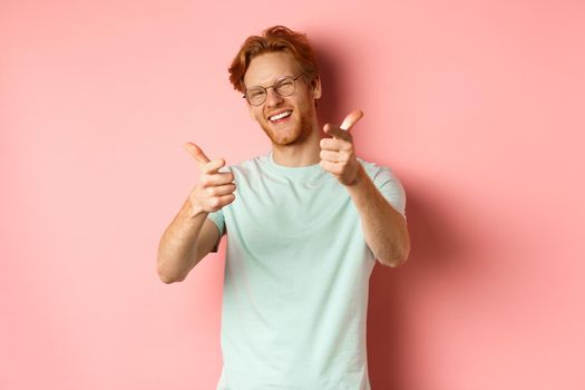 Happy young man with red hair and beard, wearing glasses, winking and smiling, pointing fingers at camera, choosing you, checking you something or congratulating winner.