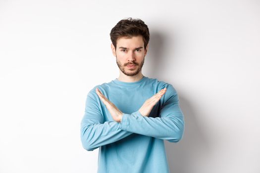 Young man with beard looking serious, making cross gesture to stop something, prohibit action, standing against white background.
