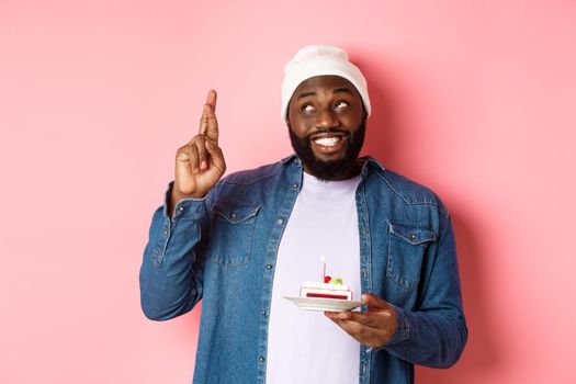 Hopeful Black guy celebrating birthday, making wish with fingers crossed, holding bday cake with candle, standing against pink background.