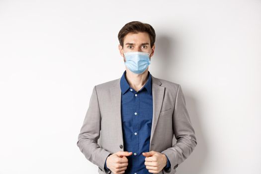Covid-19 and office workers concept. Confident businessman in medical mask and suit looking at camera, standing on white background.