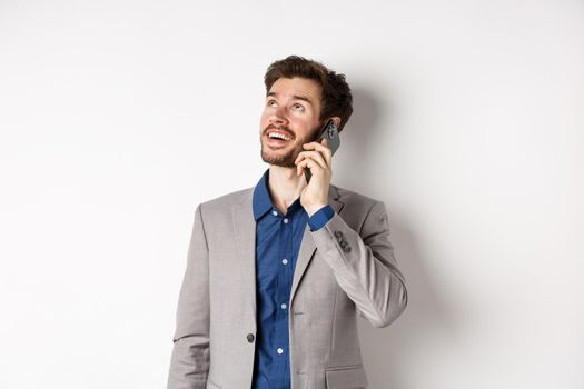 Carefree young man talking on mobile phone and smiling, looking up with dreamy face, standing in suit on white background.