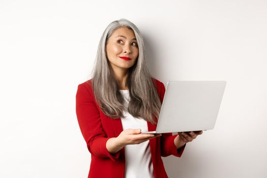 Business. Successful asian businesswoman in red blazer imaging something, working on laptop and looking upper left corner with dreamy smile, standing over white background.