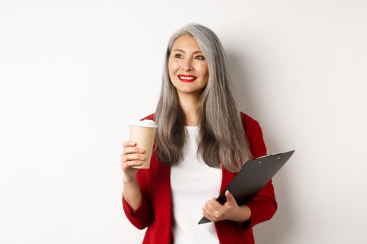 Stylish mature businesswoman in red blazer having break, holding clipboard and drinking coffee, looking left with pleased smile, white background.