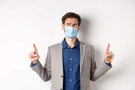 Covid-19, pandemic and business concept. Suspicious businessman in medical mask pointing up and thinking, having doubts, standing on white background.