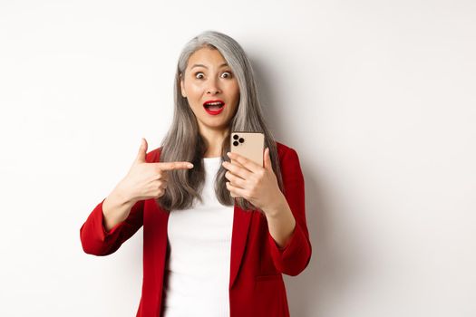 Amazed asian lady in red blazer pointing finger at smartphone, staring impressed at camera, showing something online, white background.