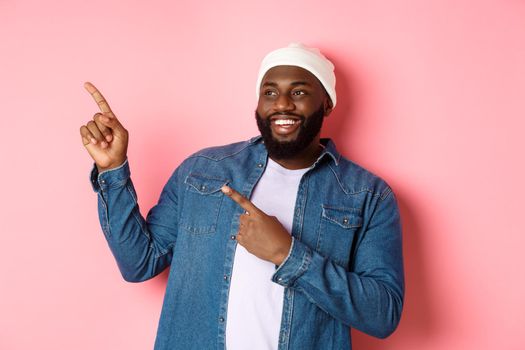 Amazed Black guy showing awesome promo offer, smiling and pointing fingers at upper left corner banner, pink background.