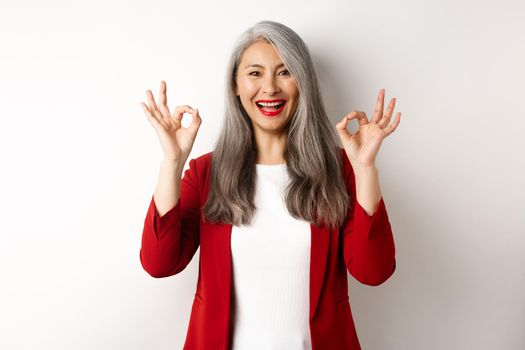 Beautiful senior asian woman in red blazer, smiling and showing okay signs, approve and say yes, standing over white background.
