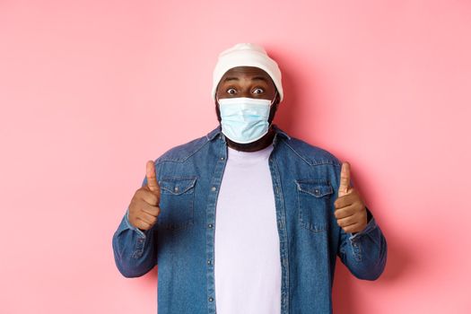 Covid-19, lifestyle and quarantine concept. Satisfied Black man in face mask showing thumbs-up, standing over pink background.