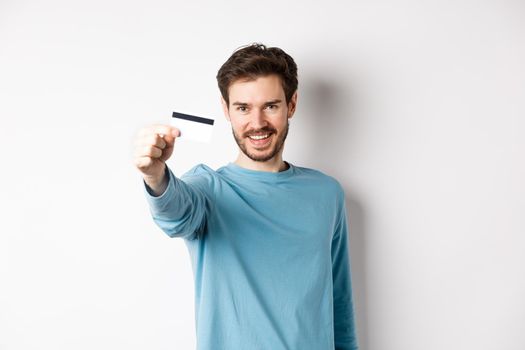 Confident young man smiling, stretch out hand and showing plastic credit card, standing on white background.
