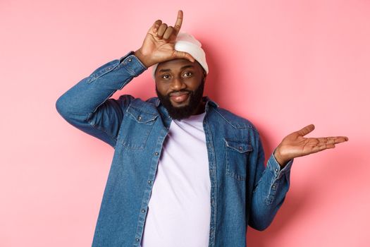 Sad african-american man showing loser sign on forehead and staring at camera, standing over pink background.