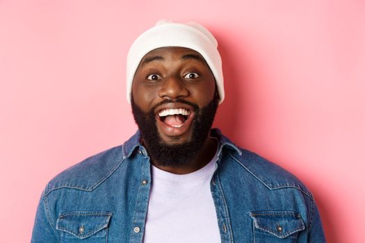 Close-up of excited bearded african-american guy in beanie staring at camera, express amazement and joy, standing over pink background.