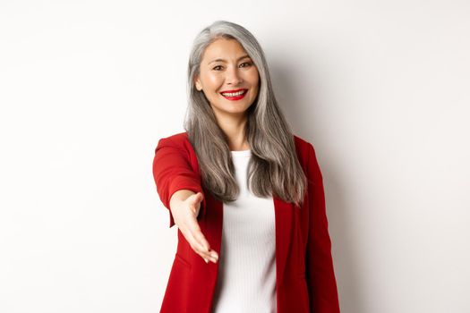 Professional asian businesswoman with grey hair, saying hello, stretch out hand for handshake and smiling, standing over white background.