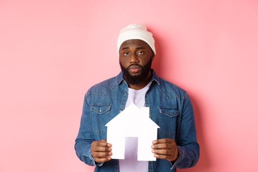 Real estate concept. Sad and tired Black man staring unamused at camera, holding paper house model, standing over pink background.