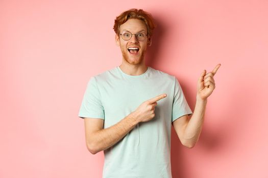 Amazed redhead man in glasses and t-shirt showing special promotion, pointing fingers at upper right corner and staring excited at camera, standing over pink background.